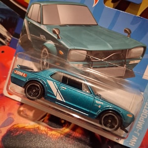 Op de afbeelding: Een turquoise Hot Wheels modelauto met witte accentstrepen en zwarte wielen. De auto zit in de originele verpakking, met het Hot Wheels logo zichtbaar. De verpakking bevat ook een afbeelding van de auto.