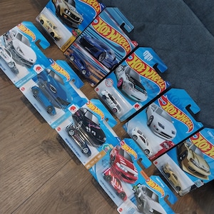 Puede incluir: Una colección de coches de juguete Hot Wheels en su embalaje original. Los coches son de varios colores, incluyendo azul, blanco, rojo y dorado. El embalaje presenta el logotipo de Hot Wheels y los nombres de los modelos de coches.