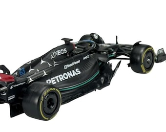 May include: A black Formula 1 race car model with Petronas and Ineos branding, next to a figurine of a driver in a black racing suit. The car has green and red accents and yellow-rimmed tires.