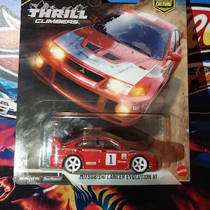 May include: A red Hot Wheels Mitsubishi Lancer Evolution VI die-cast car in its original packaging. The packaging features the "Thrill Climbers" logo and "10 Car Culture" branding. The car has white wheels and the number 1 on the side.