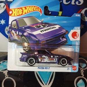 May include: A purple Hot Wheels Mazda RX-7 die-cast car. The car features white and black wheels, a white stripe, and decorative graphics. The packaging includes the Hot Wheels logo and the text "MAZDA RX-7".