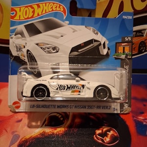 Puede incluir: Un coche de juguete Hot Wheels blanco, LB-Silhouette Works GT Nissan 35GT-RR Ver.2, en una tarjeta azul y blanca. El coche tiene ruedas negras y el logotipo de Hot Wheels. La tarjeta dice "Liberty Walk" y "154/250".