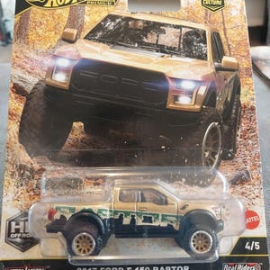 May include: A Hot Wheels Premium die-cast model of a 2017 Ford F-150 Raptor pickup truck. The truck is tan with black accents and off-road tires. The packaging features the Hot Wheels logo and the text "2017 Ford F-150 Raptor".