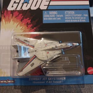 May include: A white and gray die-cast metal Combat Jet Skystriker (Grumman F-14 Tomcat) toy airplane, packaged in a clear plastic case. The packaging features the G.I. Joe logo and the text "METAL/METAL". The plane has red and blue accents.