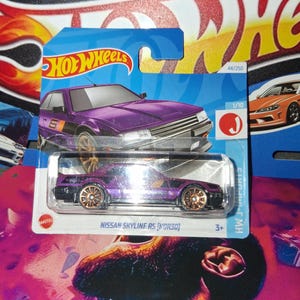 Könnte beinhalten: Ein Hot Wheels Modellauto eines lila Nissan Skyline RS (KDR30). Das Spielzeugauto hat goldfarbene Räder und ist auf einer blau-weißen Karte verpackt. Die Karte zeigt das Hot Wheels-Logo und die Nummer 44/250.
