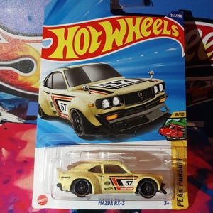 May include: A Hot Wheels die-cast model of a Mazda RX-3 in a yellow and black colour scheme. The car features the number 57 on the side and is packaged on a blue and yellow card with the Hot Wheels logo.