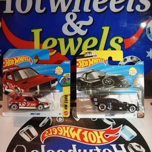 May include: Two Hot Wheels toy cars. One is red with white detailing, labelled "DRIFT BOX." The other is black, labelled "LA LIEBRE." Both are on cards with "Hot Wheels" branding and the year 2026.