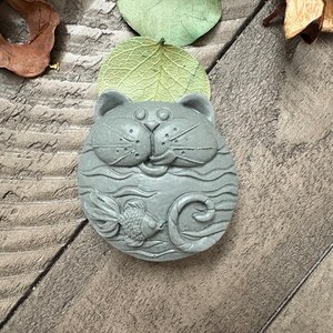 May include: A gray, cat-shaped soap with a fish design. The soap features a detailed carving of a cat's face, whiskers, and a fish swimming in wavy lines. The soap is round and has a smooth texture.
