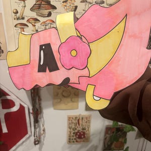 May include: A handmade, pink and yellow paper craft, possibly a mask or decorative piece, featuring a flower and the letter "A". The craft is held against a backdrop of vintage mushroom illustrations and other wall decor.