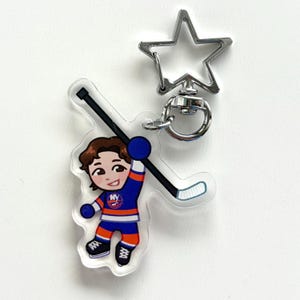 May include: An acrylic keychain featuring a cartoon hockey player wearing a blue, orange, and white jersey with an NY logo. The player holds a black hockey stick. The keychain includes a silver-toned metal star-shaped clasp and a circular ring.