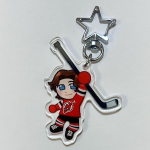 Puede incluir: Un llavero acrílico transparente con un jugador de hockey de dibujos animados con una camiseta roja con detalles en negro y blanco. El llavero tiene un cierre plateado en forma de estrella y un palo de hockey.