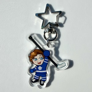 May include: A clear acrylic keychain featuring a cartoon hockey player in a blue uniform with a white maple leaf logo. The keychain has a silver star-shaped clasp and a hockey stick.