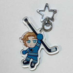 May include: A clear acrylic keychain featuring a cartoon hockey player in a blue and white uniform, holding a hockey stick. The keychain has a silver star-shaped clasp.