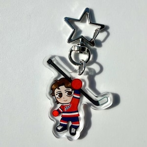 Op de afbeelding: Een heldere acryl sleutelhanger met een cartoon hockeyspeler in een rood, wit en blauw uniform, die een hockeystick vasthoudt. De sleutelhanger heeft een zilveren stervormige sluiting.