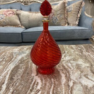 Puede incluir: Una botella de vidrio roja con un diseño de espiral y un tapón en forma de hoja. La botella está sentada sobre una mesa de mármol.