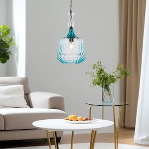 May include: A teal blue glass pendant light fixture with a clear glass shade and a black cord. The light is hanging over a white coffee table with a bowl of fruit on it.