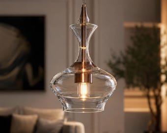 Puede incluir: Una lámpara colgante con una pantalla de cristal transparente en forma de campana, con un vástago central y un casquillo en tono cobre metálico. La lámpara está suspendida por una fina cadena metálica y contiene una bombilla de filamento.