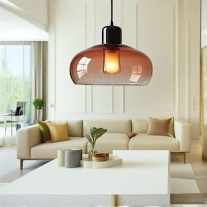 May include: A modern pendant light with a rounded, amber-colored glass shade and a black fixture. The light hangs from a black cord and illuminates a living room with a white sofa and coffee table.