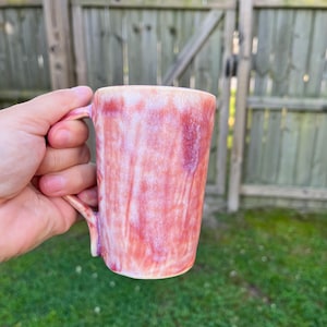 Puede incluir: Una taza de cerámica hecha a mano con un diseño veteado único en tonos rojos y rosas. La taza tiene un asa curvada y un acabado liso y brillante. La taza se sostiene en una mano, con un fondo borroso de una valla de madera y hierba verde.