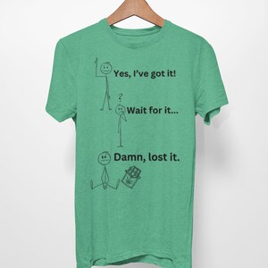 May include: A green t-shirt featuring a stick figure illustration showing the stages of losing something. The text reads "Yes, I've got it!"  "Wait for it..." and "Damn, lost it."