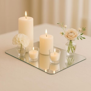 May include: A display of lit candles and small floral arrangements on a mirrored tray. The arrangement includes two pillar candles, two votive candles, and two small vases with flowers. The candles are ivory, and the flowers are light pink and white.