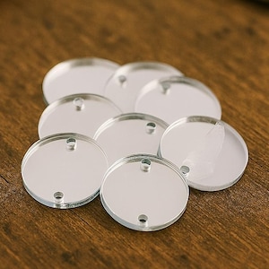 Op de afbeelding: Een verzameling kleine, heldere, cirkelvormige acryl blanks met twee gaten om te knutselen. De blanks zijn reflecterend en liggen op een houten oppervlak. Een houten deuvel, een garenklos en een schaar staan op de achtergrond.