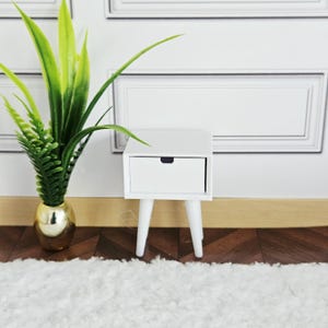 Op de afbeelding: Een kleine, witte nachtkastje met een enkele lade en drie schuine poten. Het nachtkastje staat op een donkere houten vloer, naast een gouden vaas met een groene plant. De achtergrond is een witte muur met decoratieve lijstwerk.