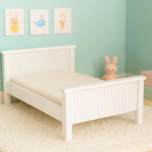 Puede incluir: Una cama de muñecas de madera blanca con un colchón descansa sobre una alfombra blanca y esponjosa. La cama tiene un cabecero y un piecero de listones. Obras de arte en colores pastel y una figura de conejito rosa decoran la habitación, creando un tema de guardería.
