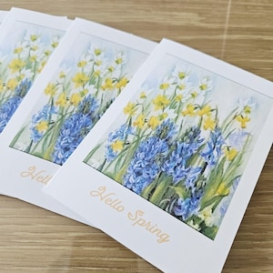 Op de afbeelding: Verschillende witte kaarten met bloemkunst met blauwe hyacinten, gele narcissen en witte bloemen. De woorden "Hello Spring" zijn in lichtbruin script op de onderkant van de kaarten gedrukt. De kaarten zijn op een houten oppervlak gestapeld.