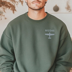 Può includere: Felpa girocollo verde militare con una piccola grafica di aereo e la scritta "N12345". La felpa è di colore pieno e ha una vestibilità rilassata. L'immagine mostra anche una persona che indossa la felpa.