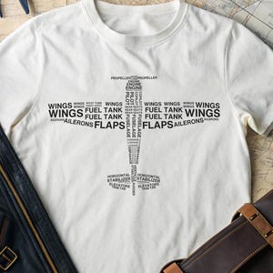 以下が含まれることがあります： 飛行機の部品を説明する言葉（翼、燃料タンク、フラップ、エルロンなど）で構成された、黒い飛行機のデザインが施された白いTシャツ。