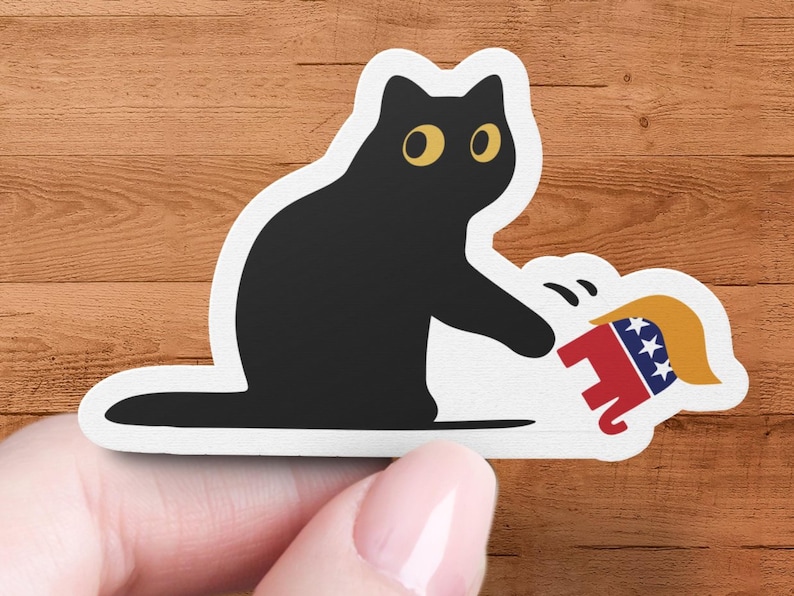 Puede incluir: Una pegatina de gato negro con ojos amarillos, sosteniendo un elefante de dibujos animados con un dise&ntilde;o rojo, blanco y azul. El elefante tiene un peinado rubio. La pegatina est&aacute; sobre una superficie de madera.