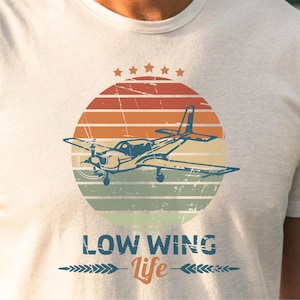 May include: A white t-shirt with a vintage-style graphic of a small airplane flying in front of a sunset. The text "LOW WING Life" is printed below the graphic.