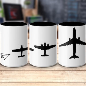 May include: White ceramic mug with a black interior and handle, featuring a progression of airplane silhouettes. The design starts with a paper airplane, transitioning to propeller planes, and ending with a jet. The mug is set on a wooden surface.