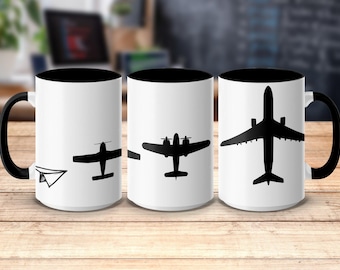 May include: White ceramic mug with a black interior and handle, featuring a progression of airplane silhouettes. The design starts with a paper airplane, transitioning to propeller planes, and ending with a jet. The mug is set on a wooden surface.