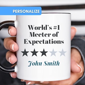 Può includere: Una tazza in ceramica bianca con manico e bordo neri. La parte anteriore presenta il testo nero "World's #1 Meeter of Expectations" sopra tre stelle nere piene e due stelle grigio chiaro. Il nome "John Smith" è stampato in corsivo verde acqua.