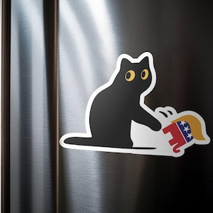Puede incluir: Una pegatina de gato negro con ojos amarillos, sosteniendo un elefante de dibujos animados con un dise&ntilde;o rojo, blanco y azul. La pegatina est&aacute; en la puerta plateada de una nevera.
