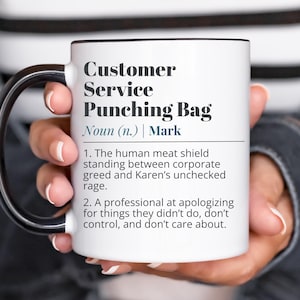 May include: White ceramic mug with a black handle and rim, featuring the text "Customer Service Punching Bag" and a humorous definition. The mug is held by a person. A fun gift for customer service professionals.