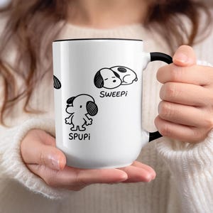 Può includere: Tazza in ceramica bianca con manico e bordo neri, con illustrazioni di cani dei cartoni animati in bianco e nero. La tazza ha le parole "SWEEPI" e "SPUPI" stampate.