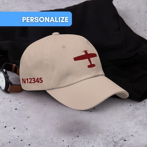 Op de afbeelding: Beige baseballpet met een rode vliegtuigafbeelding en de tekst "N12345". Het woord "PERSONALISEER" staat in een blauwe rechthoek boven de pet. Een horloge en een zwart kledingstuk staan op de achtergrond.