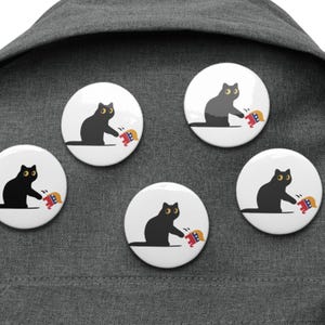 Könnte beinhalten: Fünf weiße runde Ansteckbuttons mit einer Illustration einer schwarzen Katze, die nach einem kleinen, farbenfrohen Objekt greift. Die Buttons sind an einem grauen Rucksack befestigt. Die Katze hat gelbe Augen.