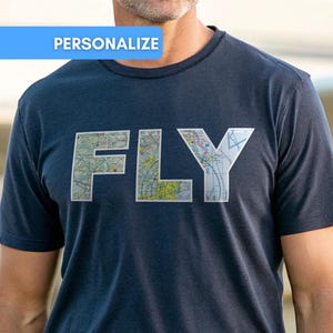 May include: A dark navy blue short-sleeved t-shirt featuring the word FLY printed in large block letters across the chest. The letters are filled with a detailed multicoloured aeronautical map pattern. A blue banner with the text PERSONALIZE is visible.