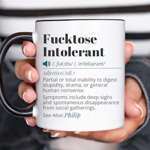 Puede incluir: Una taza de cerámica blanca con asa y borde negros, que presenta el texto negro "Fucktose Intolerant" seguido de una pronunciación fonética, una definición sobre la incapacidad de digerir la estupidez y una referencia "See Also: Philip".