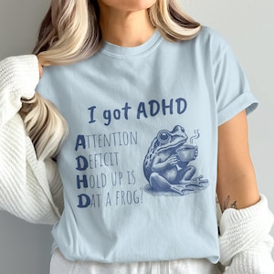 Pode incluir: Camiseta azul claro com um gr&aacute;fico de sapo azul e o texto "Eu tenho TDAH. D&eacute;ficit de aten&ccedil;&atilde;o Hold Up Is Dat A Frog?"