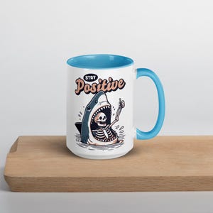 May include: White ceramic mug with a blue interior and handle. The mug features a cartoon illustration of a shark with a skeleton inside giving a thumbs up. The words "Stay Positive" are above the shark.