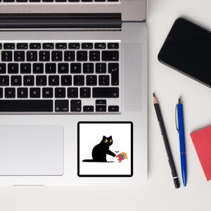 Puede incluir: Una pegatina de gato negro con ojos amarillos y un gr&aacute;fico rojo, blanco y azul. El gato est&aacute; alcanzando el gr&aacute;fico. La pegatina es cuadrada y tiene un fondo blanco. La pegatina est&aacute; sobre una superficie blanca.