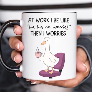 May include: A white ceramic mug with a black handle and rim featuring a cartoon illustration of a white duck with an orange beak sitting on a purple office chair while holding a small pink coffee cup. The text reads AT WORK I BE LIKE ha ha no worries THEN I WORRIES.