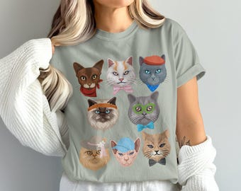 May include: A light green t-shirt with a graphic of eight cats wearing different hats and accessories. The cats are all different colors and breeds.