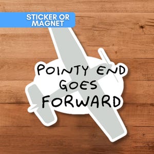 May include: A sticker or magnet featuring a stylized airplane illustration in gray and white, with the text "POINTY END GOES FORWARD" in black. A blue banner at the top reads "STICKER OR MAGNET". The design is set against a wood grain background.