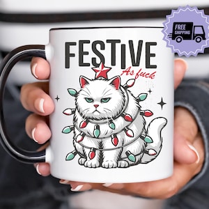 Könnte beinhalten: Weiße Keramik-Tasse mit schwarzem Henkel und Rand, mit einer grimmigen Cartoon-Katze, die in Weihnachtsbeleuchtung eingewickelt ist. Auf der Tasse steht "FESTIVE As fuck".